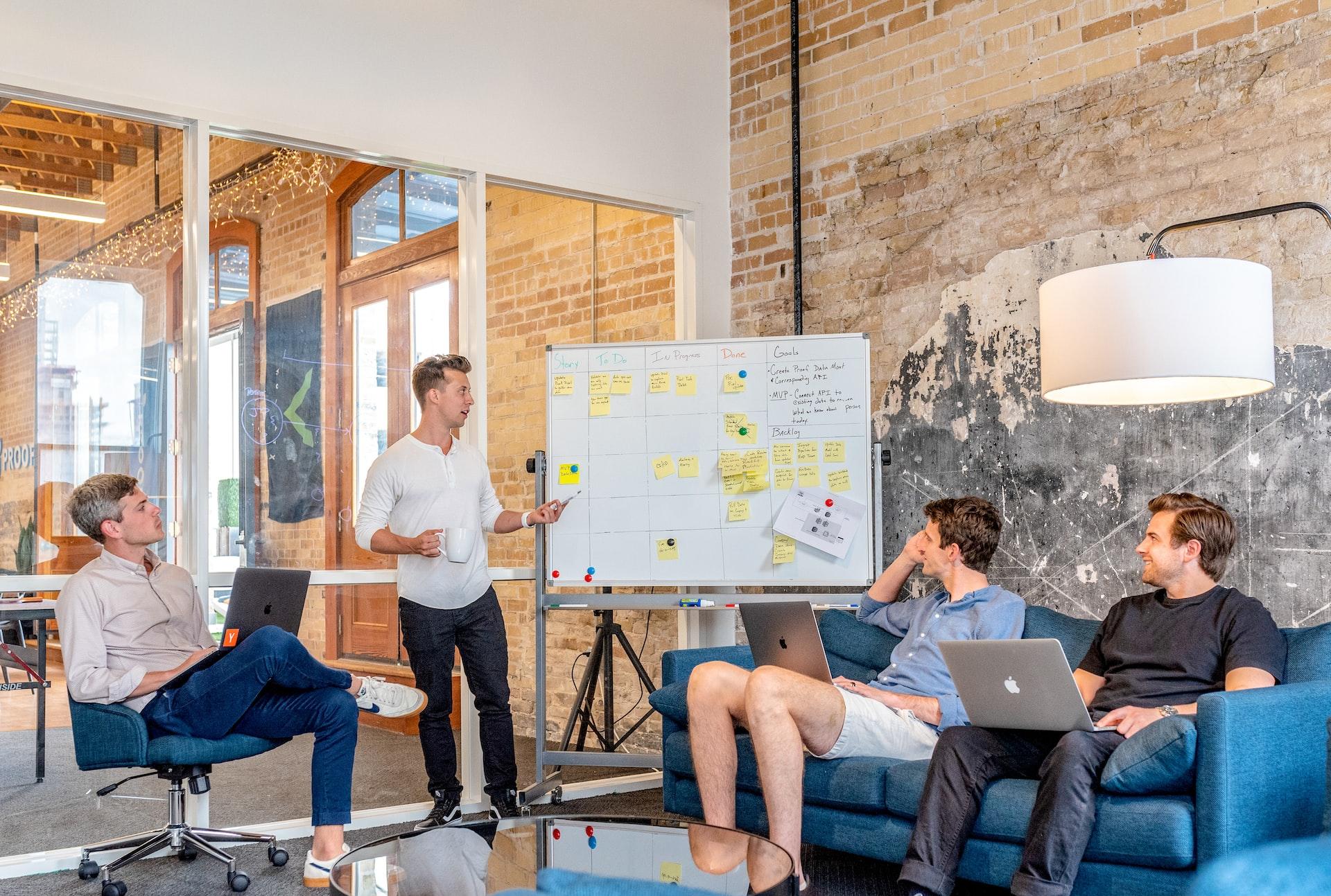 three men sitting while using laptops and watching man beside whiteboard